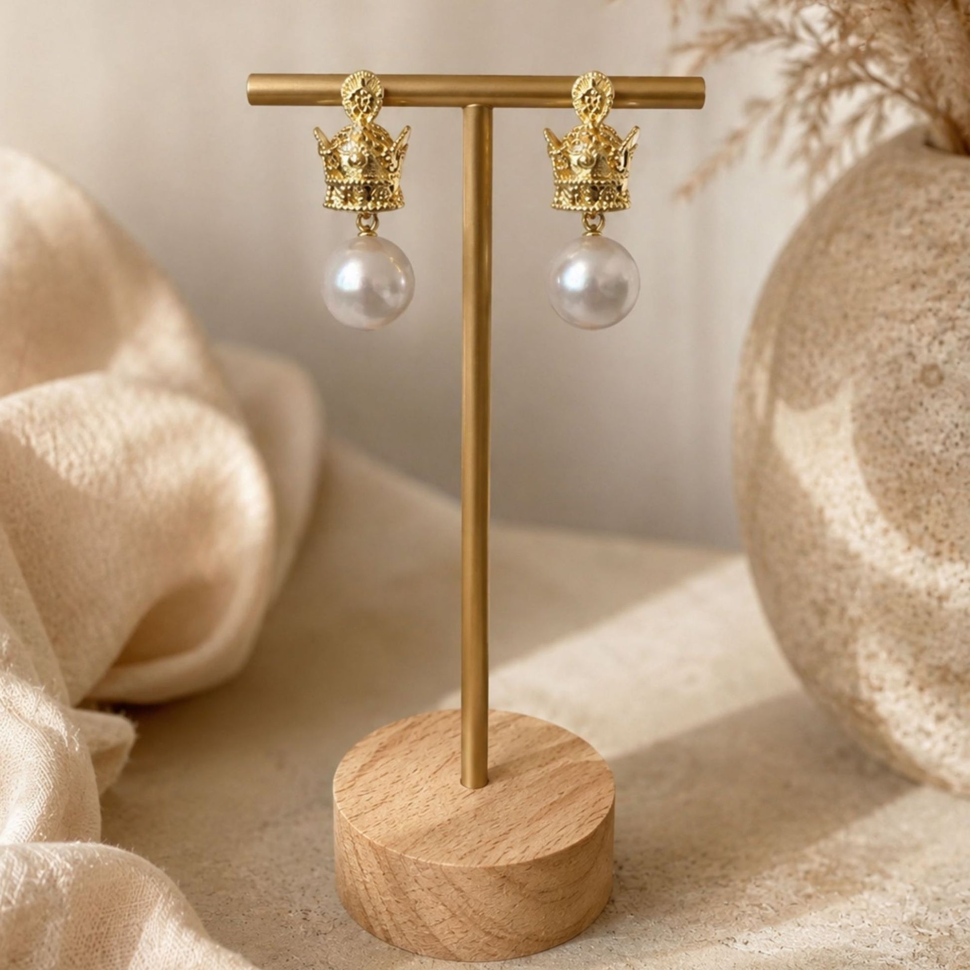 A pair of gold-tone crown drop earrings with white faux pearl pendants displayed on a wooden stand with a gold T-bar, set against a warm neutral background with a textured vase and draped fabric.