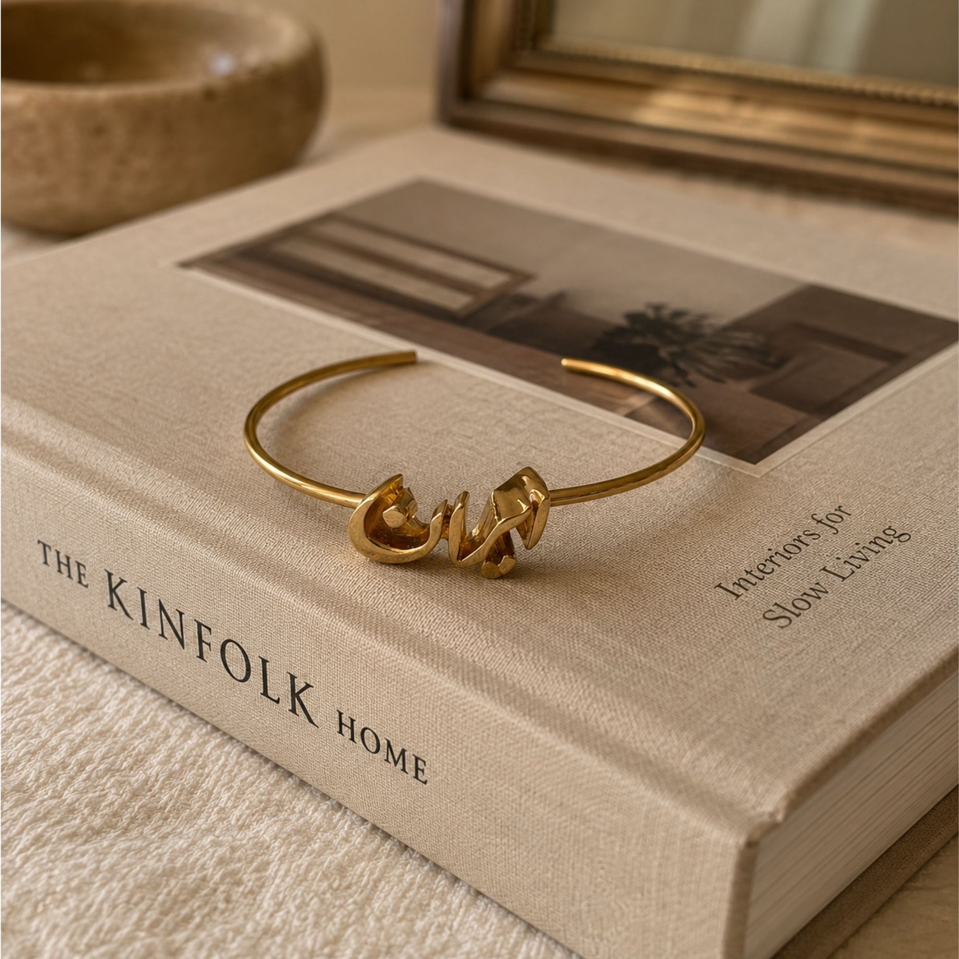 Adjustable gold-tone Iran script bangle bracelet displayed on a luxury coffee table book in an elegant lifestyle setting.