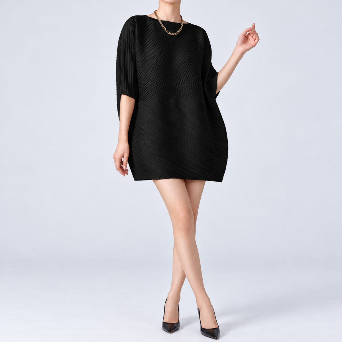 A woman modeling a black pleated mini dress with three-quarter sleeves and a relaxed silhouette, paired with black pointed-toe heels and a gold chain necklace, against a light gray studio background.
