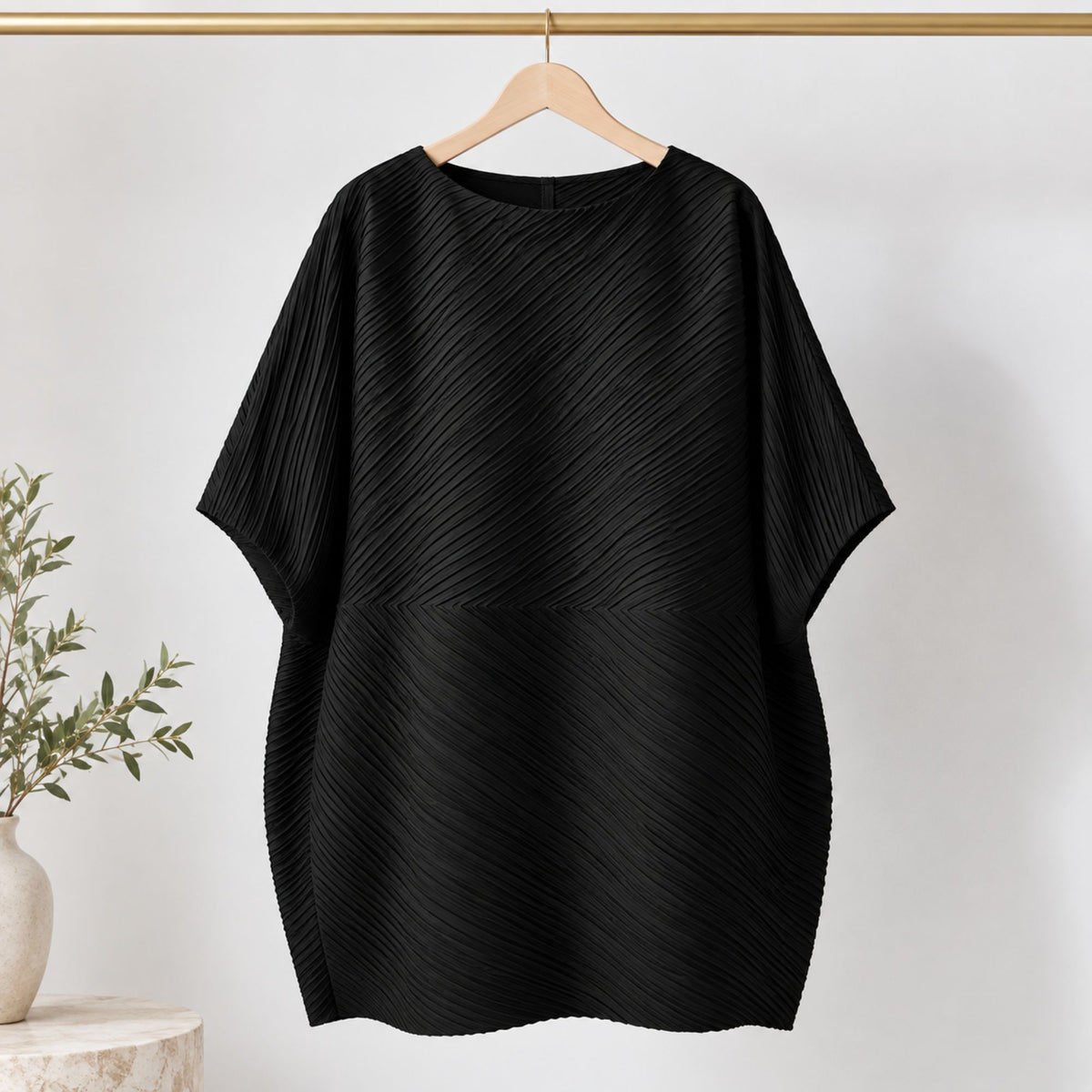 A black pleated short-sleeve top hanging on a wooden hanger against a white wall, with a small plant and vase visible at the lower left.