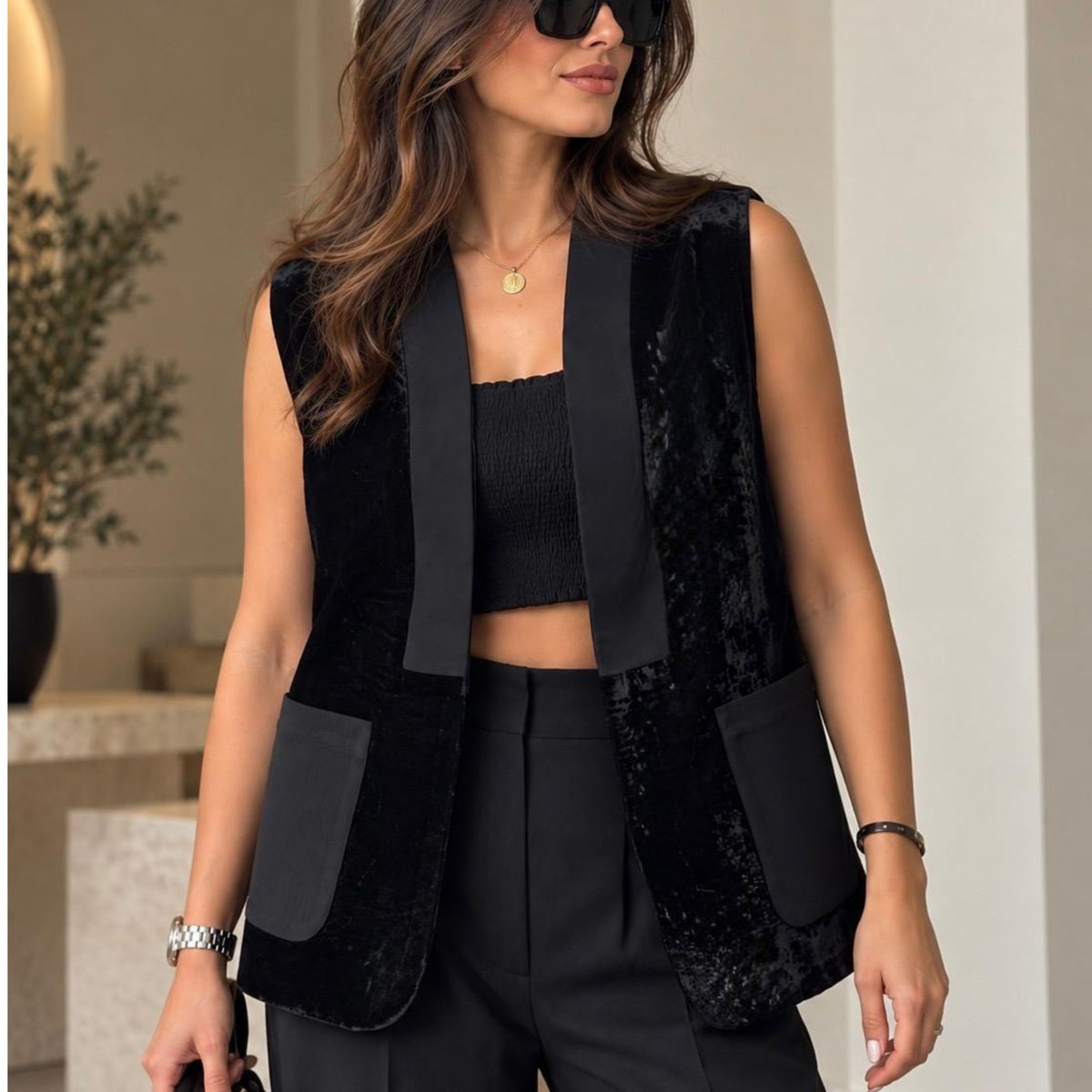 A woman wearing a sleeveless black velvet vest with satin lapels and large patch pockets, paired with a black crop top and black trousers, in a softly lit indoor setting.