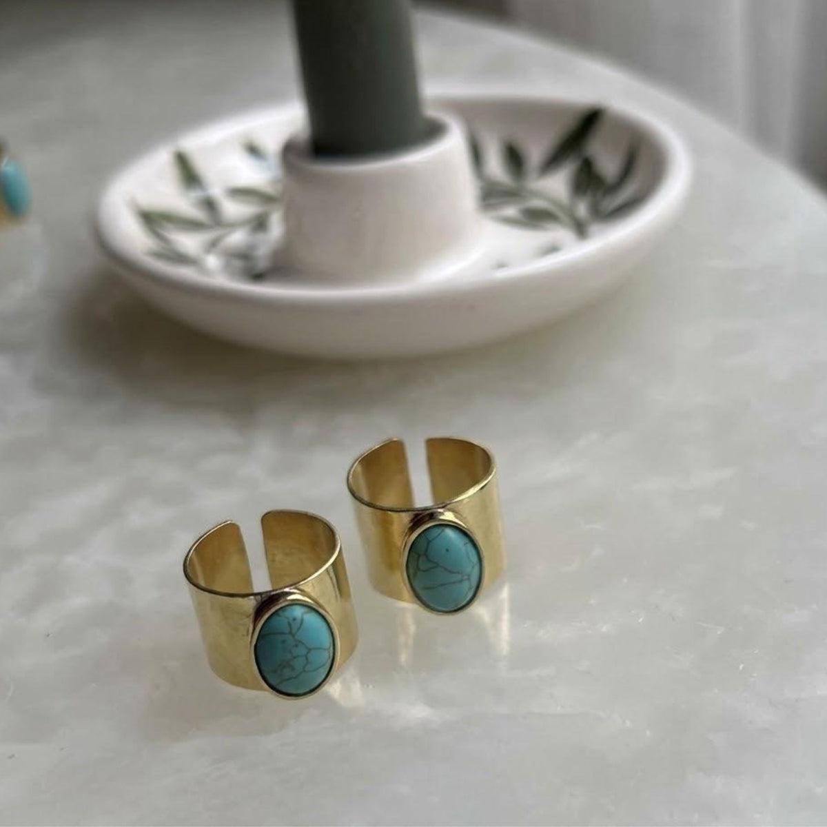Two gold-tone open-back rings with oval turquoise-look accents displayed beside a candle holder.