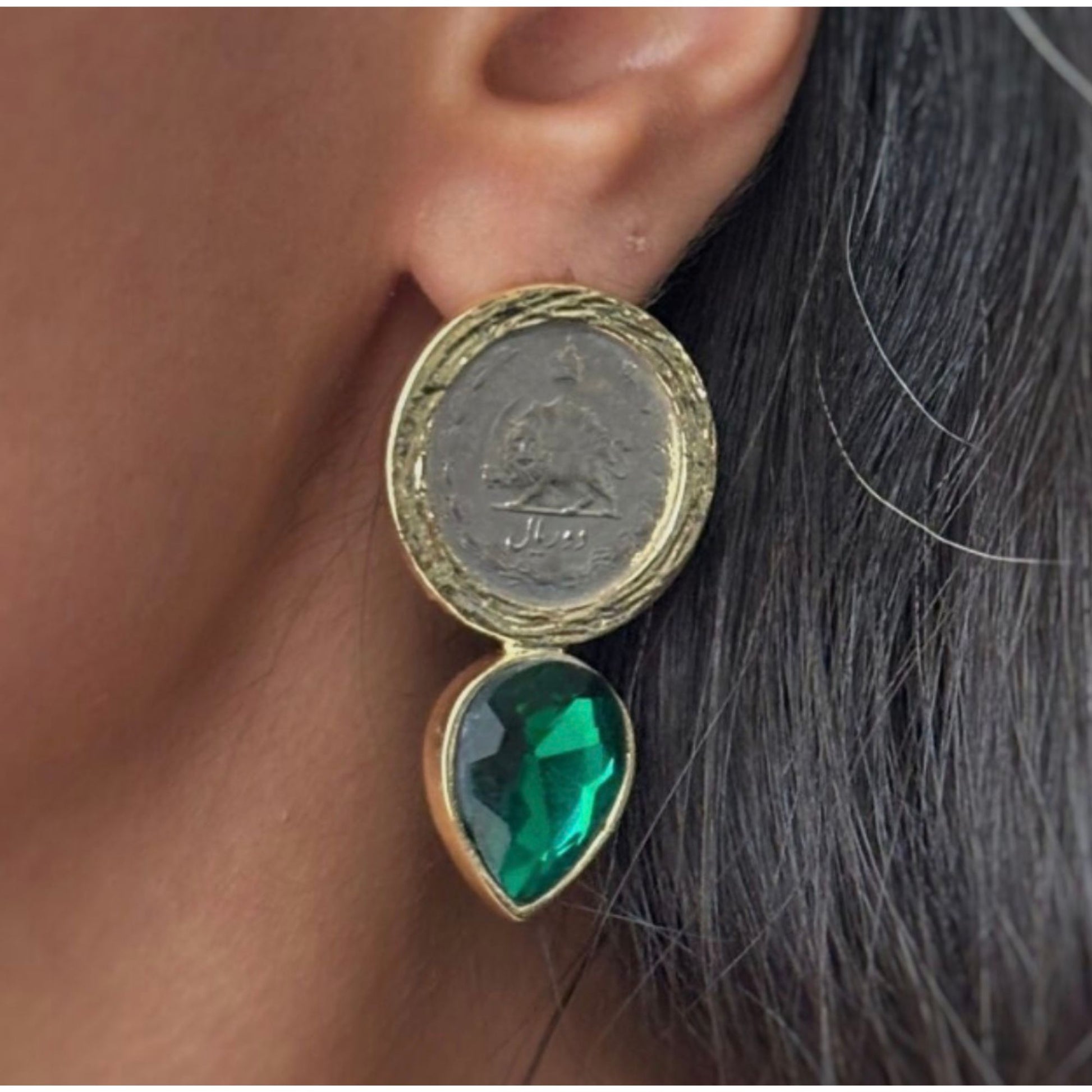 Close-up of a teardrop earring with a coin and green crystal worn by a person.