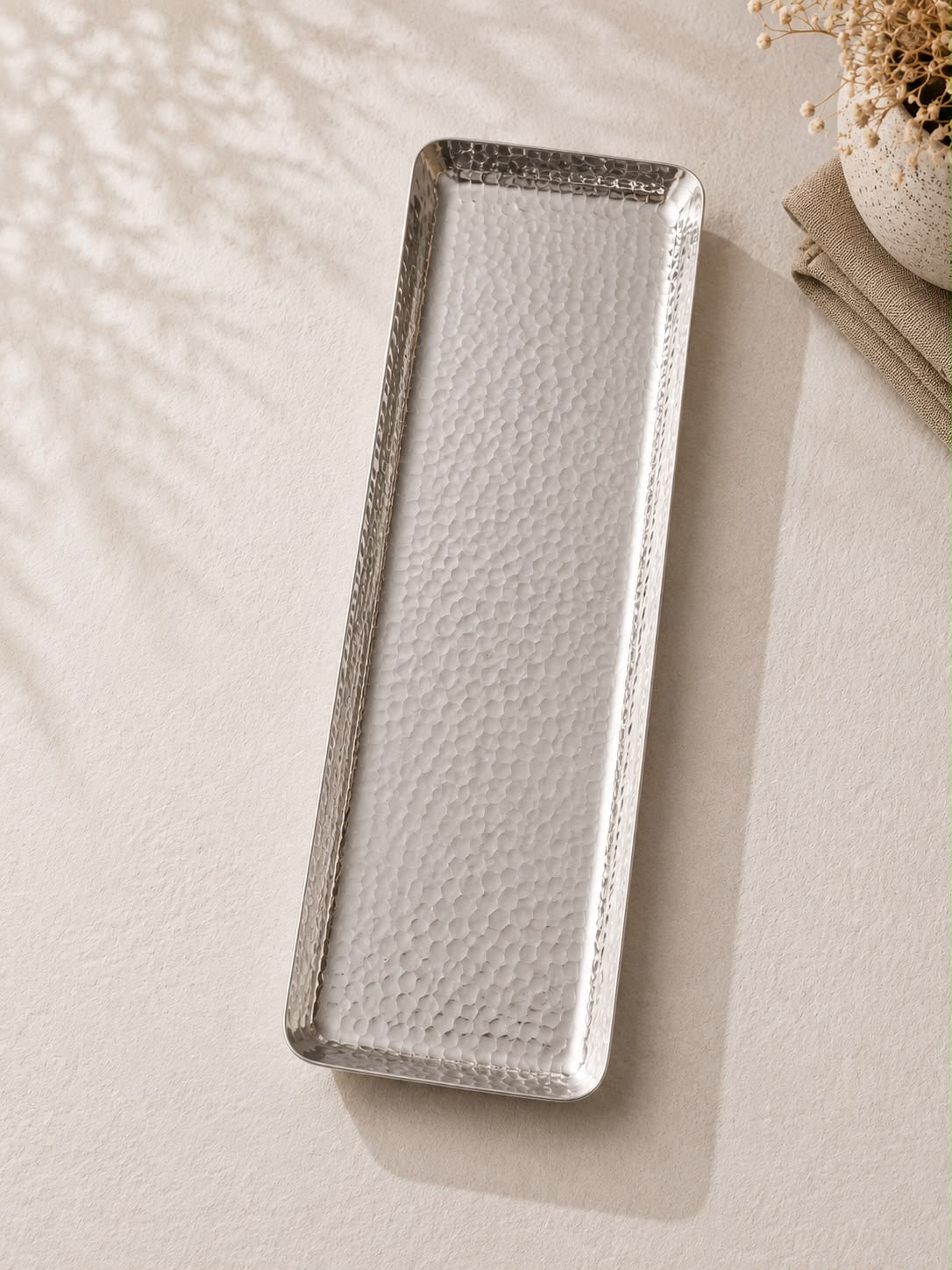 A rectangular tray with a hammered texture and beveled edges, photographed on a light beige surface with soft leaf shadows and a folded napkin nearby.