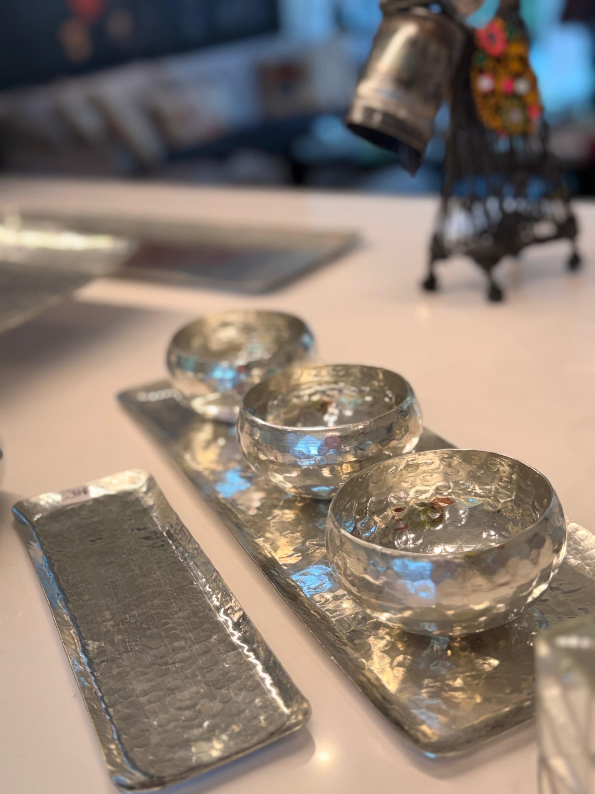 Three small hammered clear glass bowls arranged on rectangular silver trays on a white tabletop, with a softly blurred indoor background.