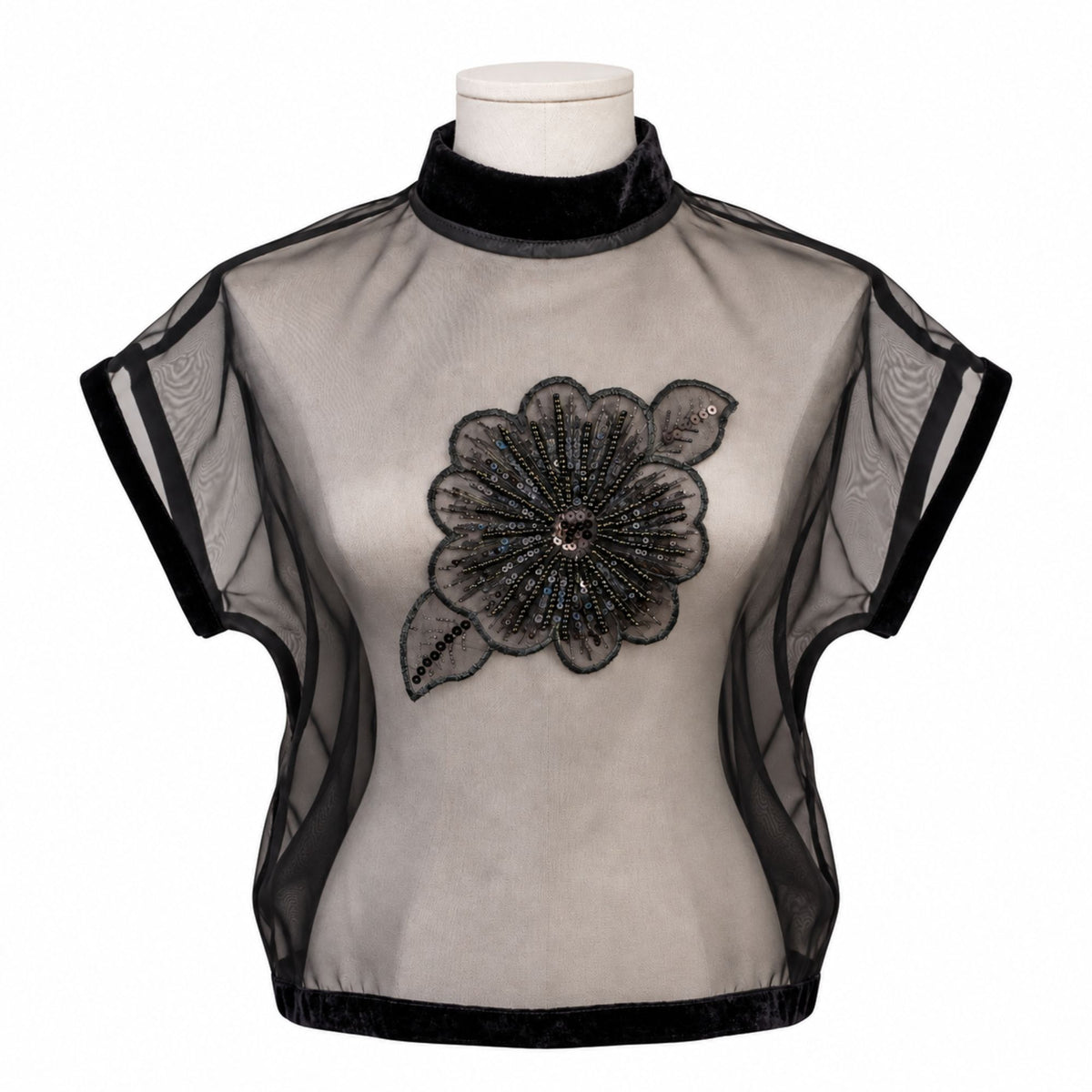 A sheer black mesh blouse with a high black velvet mock neck and a large black beaded floral applique on the chest, displayed on a mannequin torso against a white background.