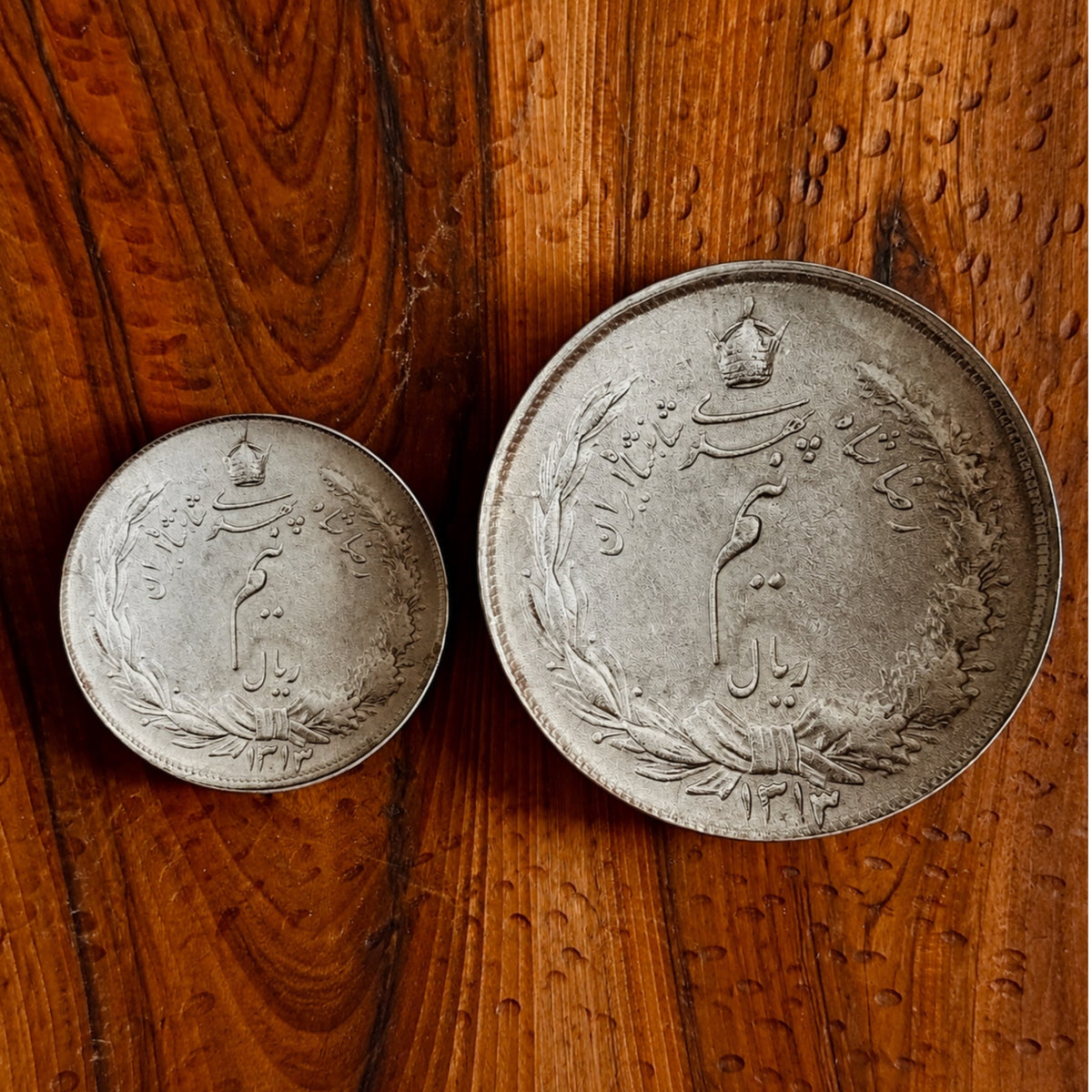 Two coins with Farsi calligraphy and a crown emblem, displayed on a rustic wooden surface.