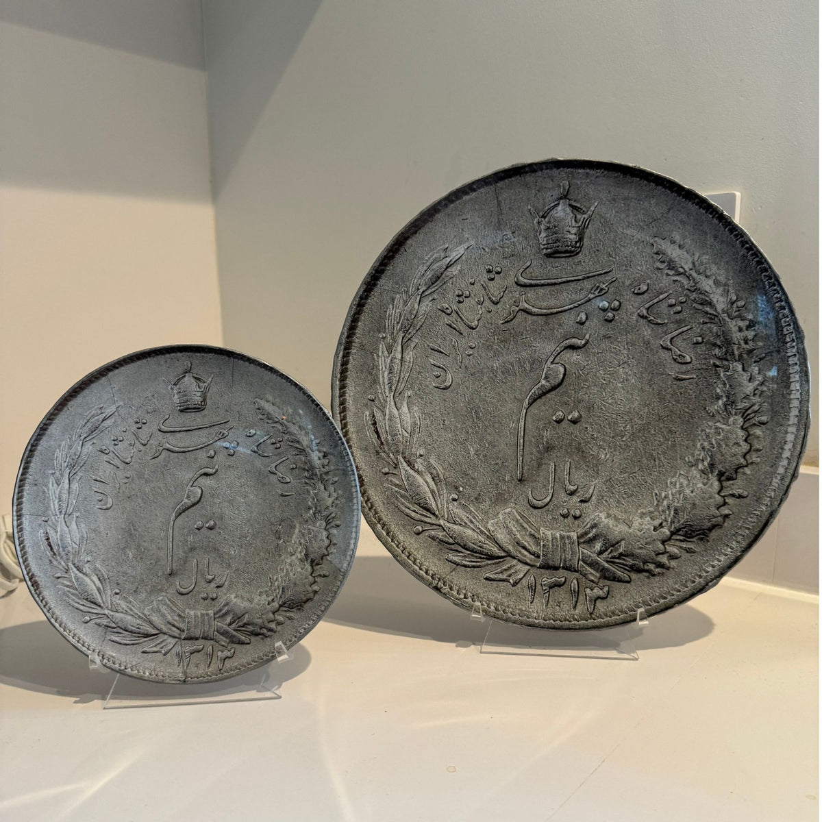 Two round coins farsi script plates in two different sizes stand against a plain light gray wall and surface.