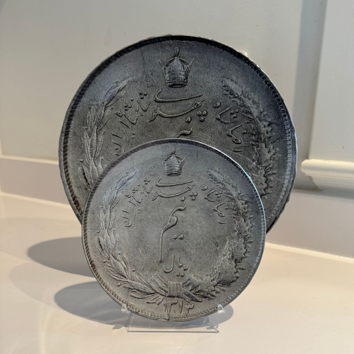 Two different sizes of plate with coin calligraphy and a crown motif, displayed on clear stands against a light gray background.