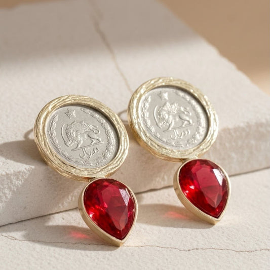 Red ruby teardrop crystals and gold-tone fashion earrings on a textured surface with pahlavi era coins shiro khorshid.