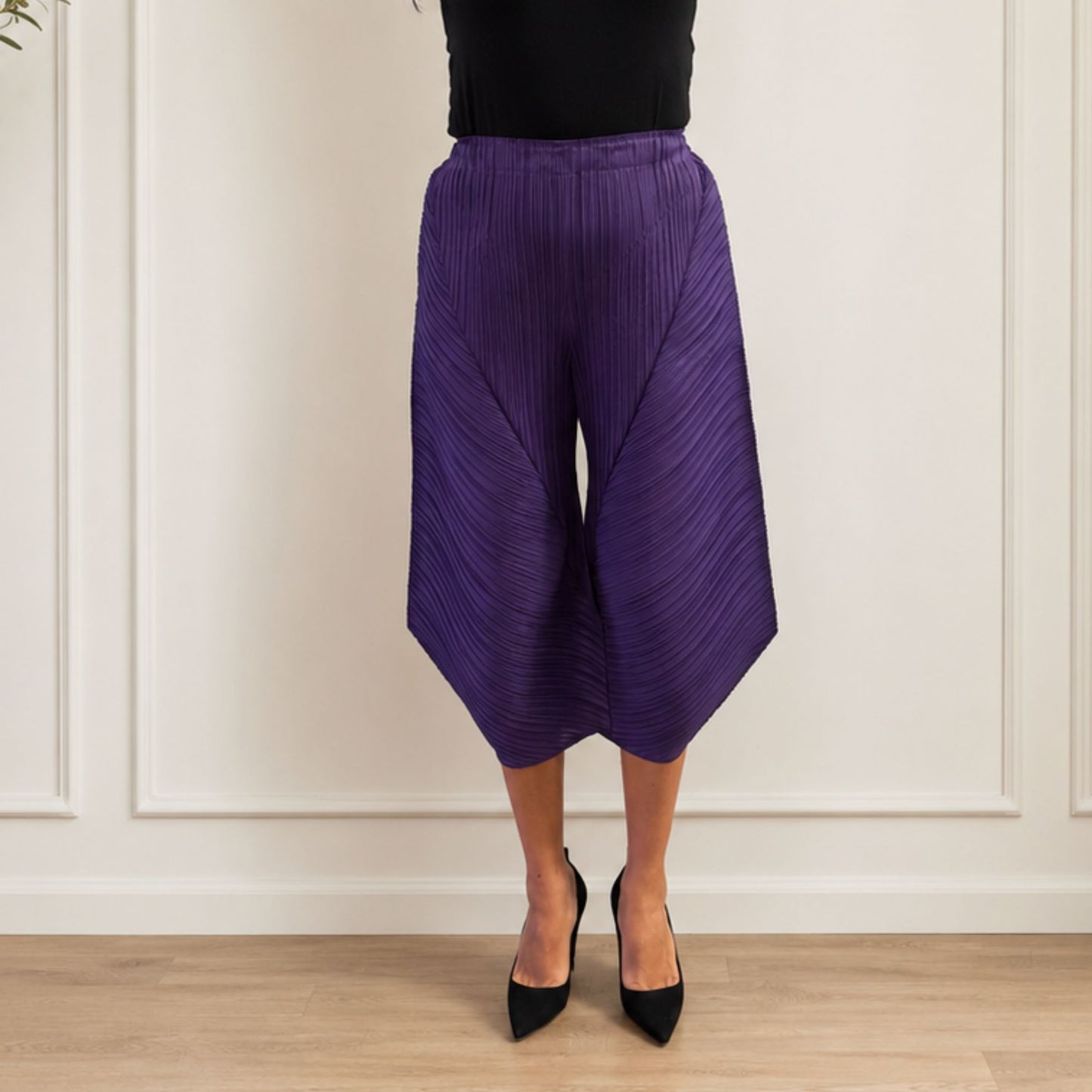 Person wearing purple pleated pants and black shoes against a white paneled wall.