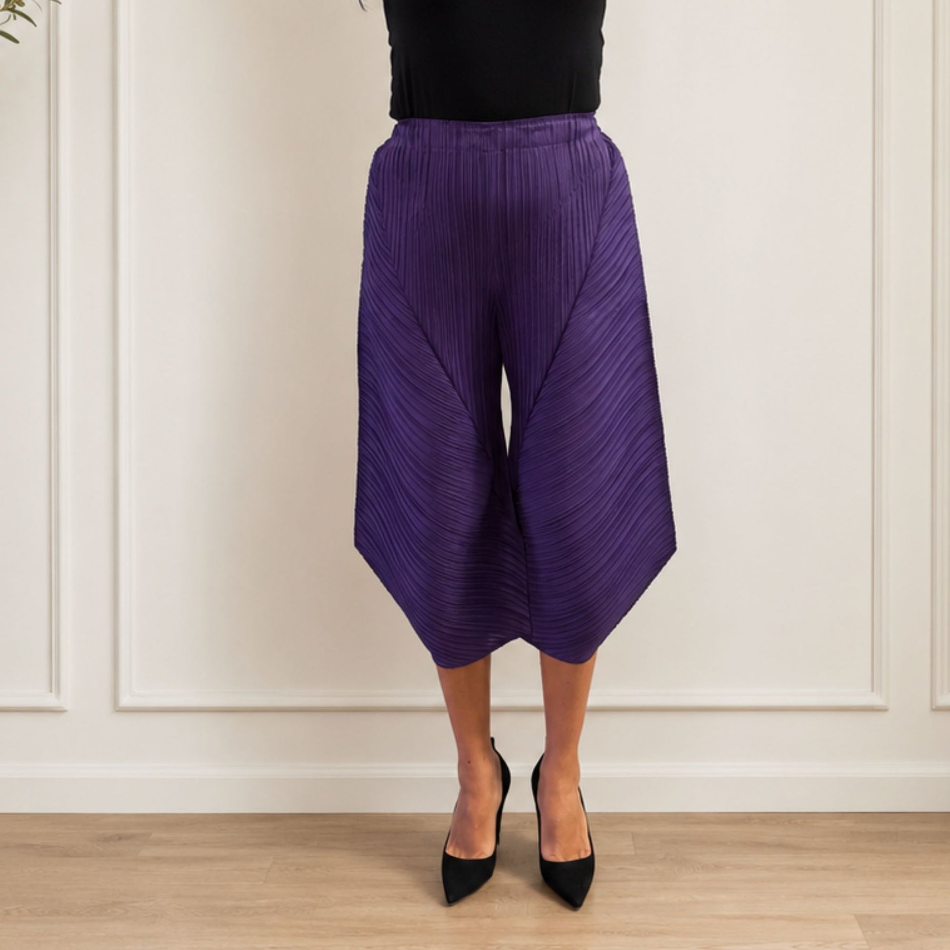 Person wearing purple pleated pants and black shoes against a white paneled wall.