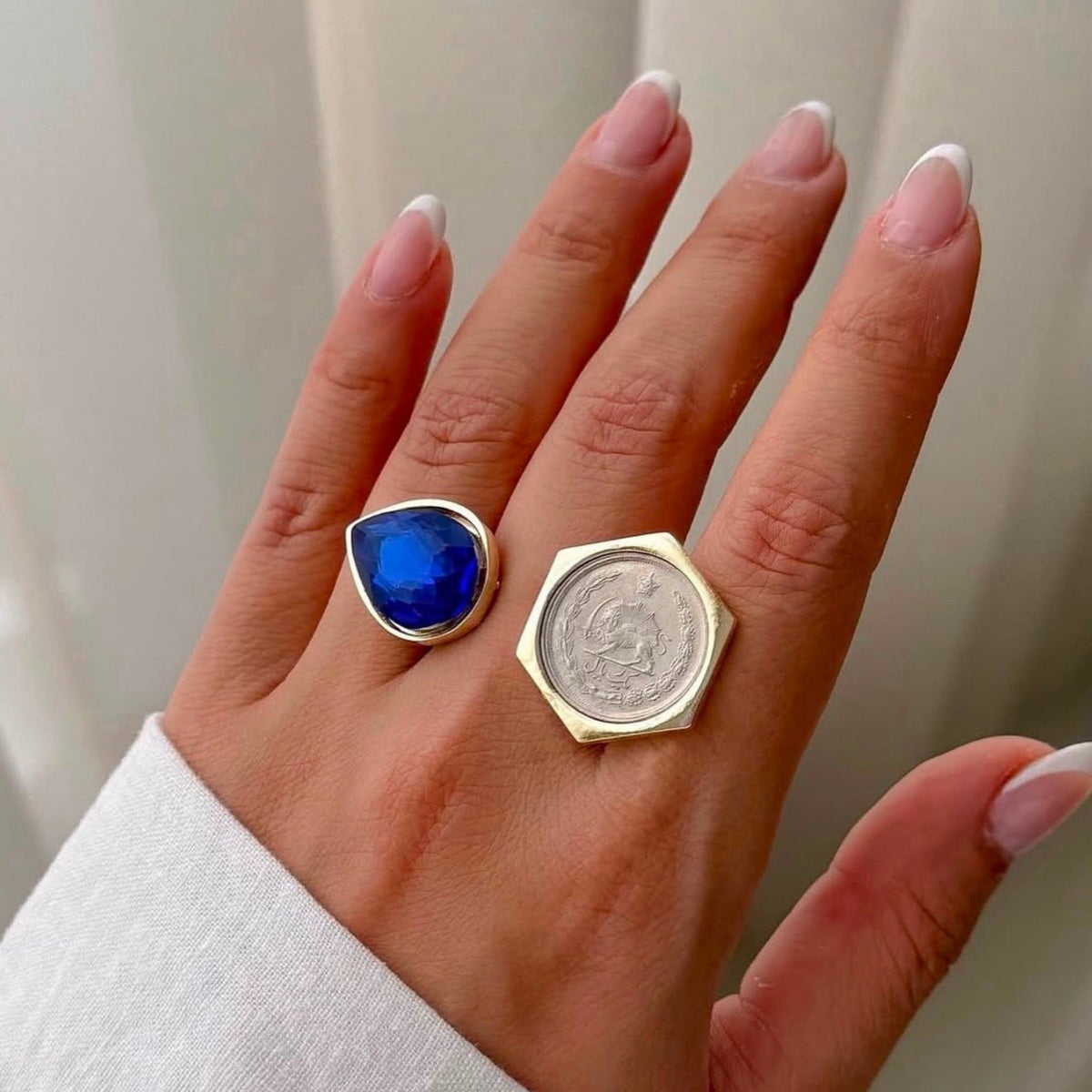 A close-up of a hand wearing an adjustable gold-tone ring with a deep blue teardrop-shaped crystal and a vintage coin ring, shown against a soft light background.