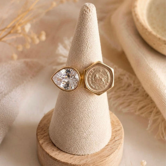 A gold-tone ring displayed on a beige fabric cone stand, featuring a marquise-cut clear crystal on one side and a hexagonal coin on the other, styled with dried grasses on a neutral beige background.