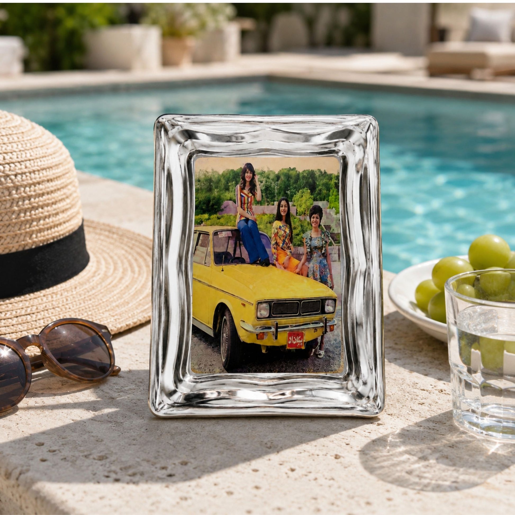 Vintage mini art and serving tray featuring a classic yellow Peykan design inspired by the nostalgia of old Iran. Polished metal tray, 7 × 5 inches styled on a stone patio next to a straw hat, sunglasses, a glass of water, and green grapes, with a swimming pool in the background.